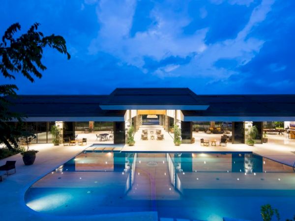 An outdoor hotel pool at dusk, lit by lights, with lounge chairs and a covered building under a blue sky.