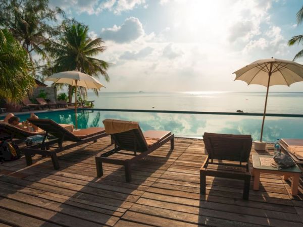 A beachside resort deck with wooden loungers, umbrellas, and calm ocean waves under a partly cloudy sky.