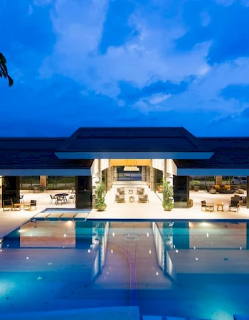 A modern outdoor resort with a lit swimming pool, lounge chairs, and palm trees under a vivid blue evening sky.