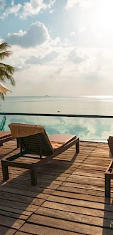 Beachside resort deck with wooden lounge chairs, umbrellas, and a pool overlooking the calm ocean under cloudy skies.