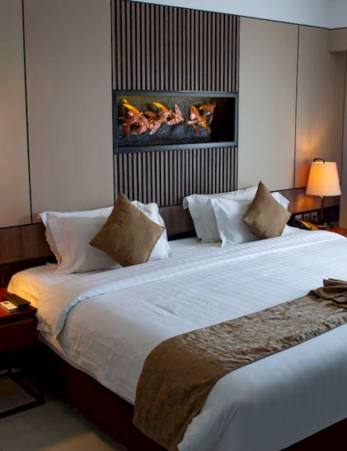 A modern hotel bedroom with a neatly made bed, bedside lamps, curtains, and a built-in fireplace with burning logs.