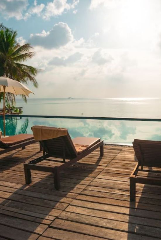 A tropical beach resort deck with sun loungers and umbrellas facing the calm ocean under a partly cloudy sky.