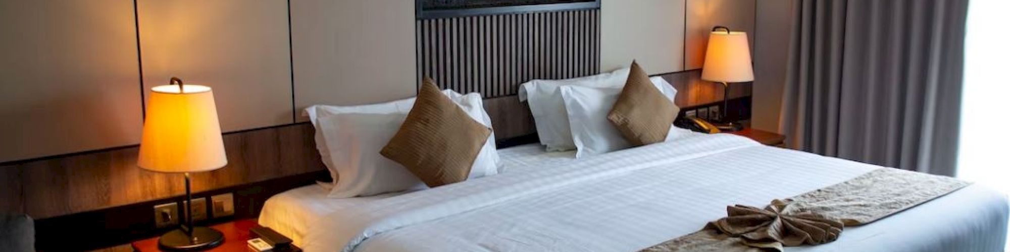 A modern hotel room with a neatly made bed, bedside lamps, and a wall fireplace featuring glowing logs in the back.