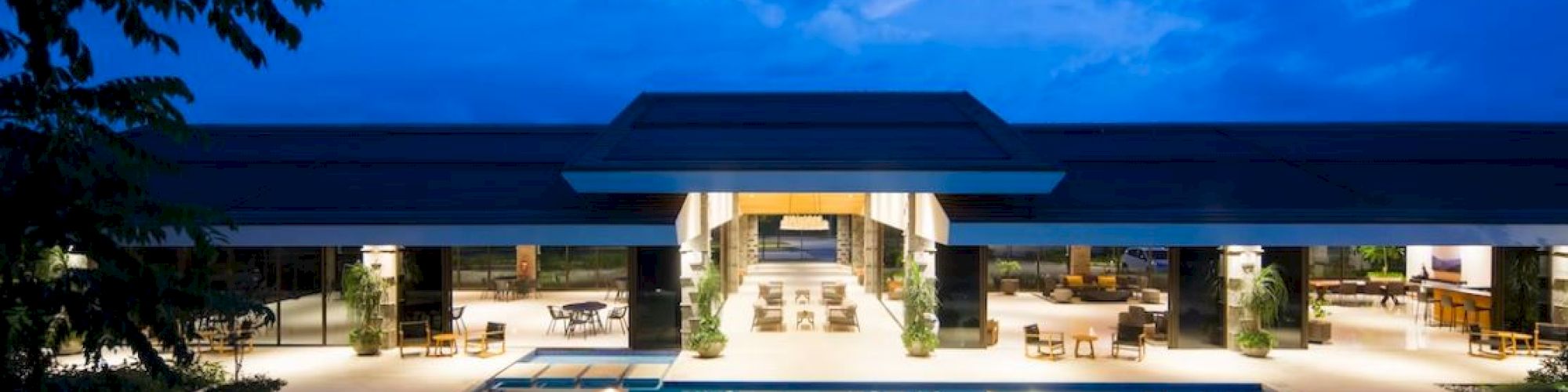 A modern hotel courtyard with a lit swimming pool, outdoor seating, and a dramatic blue evening sky.