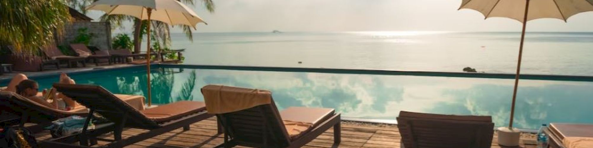 A beachfront resort deck with lounge chairs, umbrellas, and a pool facing the calm ocean under a partly cloudy sky.