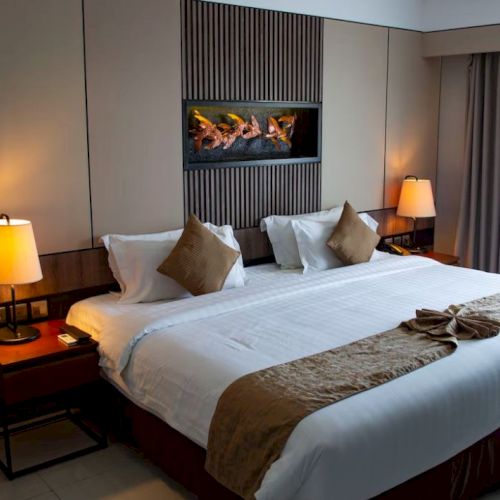 A cozy hotel room with a neatly made bed, pillows, bedside lamps, and a wall fireplace with glowing logs.