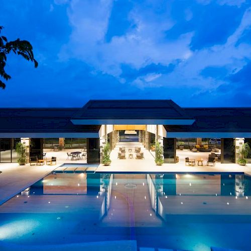 A modern resort courtyard with a lit swimming pool, lounge chairs, and a sleek building under a vivid blue evening sky.