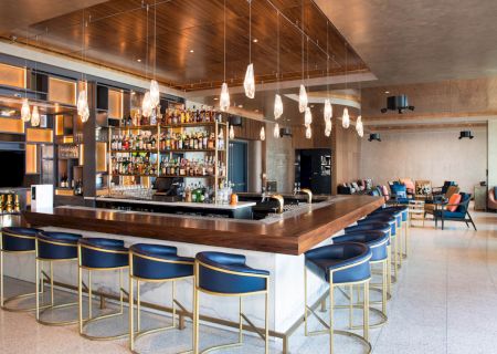 A modern hotel bar with a polished wooden counter, glowing bottles on shelves, pendant lights, and blue bar stools.