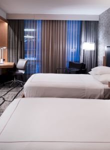 Modern hotel room with two beds, wall-mounted TV, desk, and bedside lamps beside window curtains.