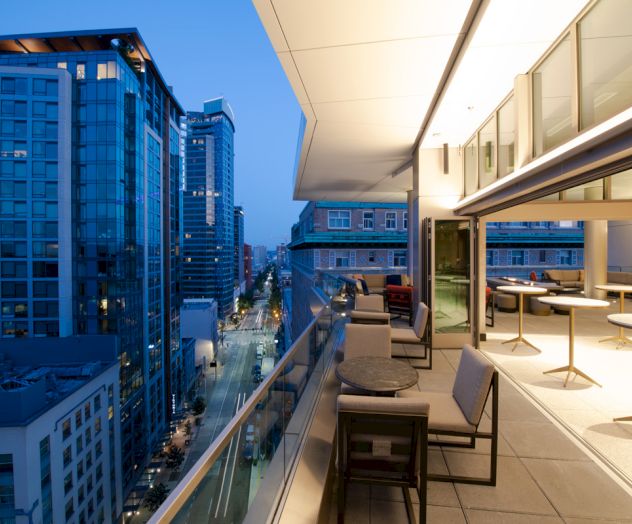 Balcony terrace with lounge chairs overlooking a downtown street and skyscrapers at dusk.