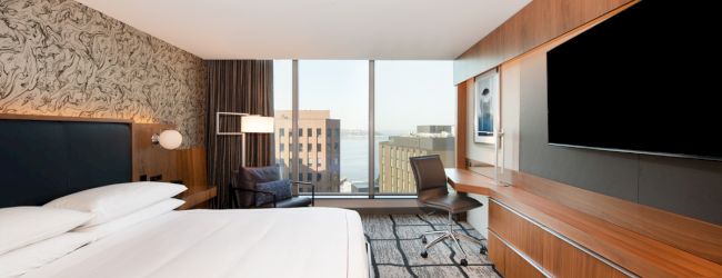 Modern hotel room with a large bed, window view, desk chair, and flat-screen TVs.