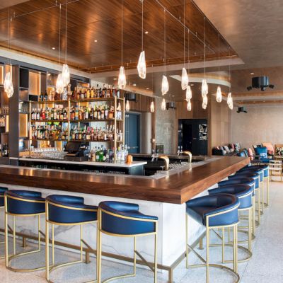 Modern hotel bar with a marble counter, blue stools, hanging pendant lights, stocked bottle shelves, and lounge seating.