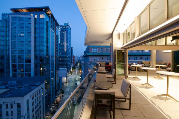A modern rooftop terrace with lounge chairs and tables overlooking a city of tall buildings at dusk.