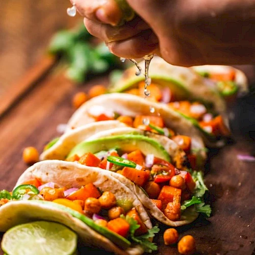 A person pours taco filling and sauce onto tortillas loaded with vegetables, herbs, and beans on a wooden table with lime.