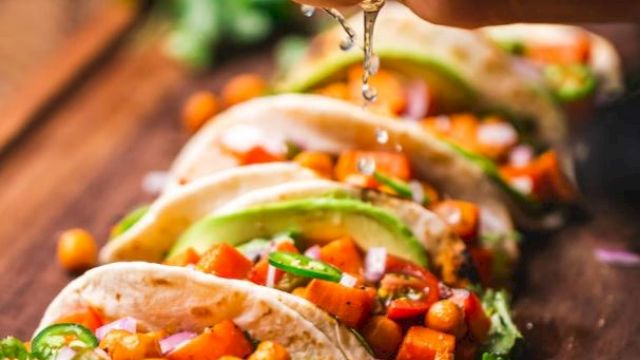 A person pours taco filling and sauce onto tortillas loaded with vegetables, herbs, and beans on a wooden table with lime.
