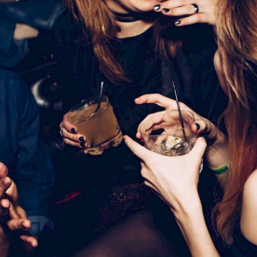 Two people at a dark party sip cocktails through straws, with hands and rings visible under moody lighting.
