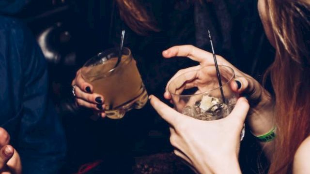 Two people at a dark party sip cocktails through straws, with hands and rings visible under moody lighting.