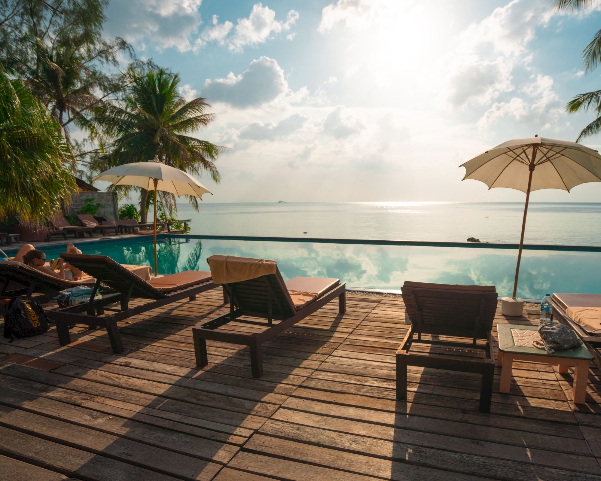 Tropical resort deck with a pool, lounge chairs and umbrellas overlooking the ocean under sunny clouds.