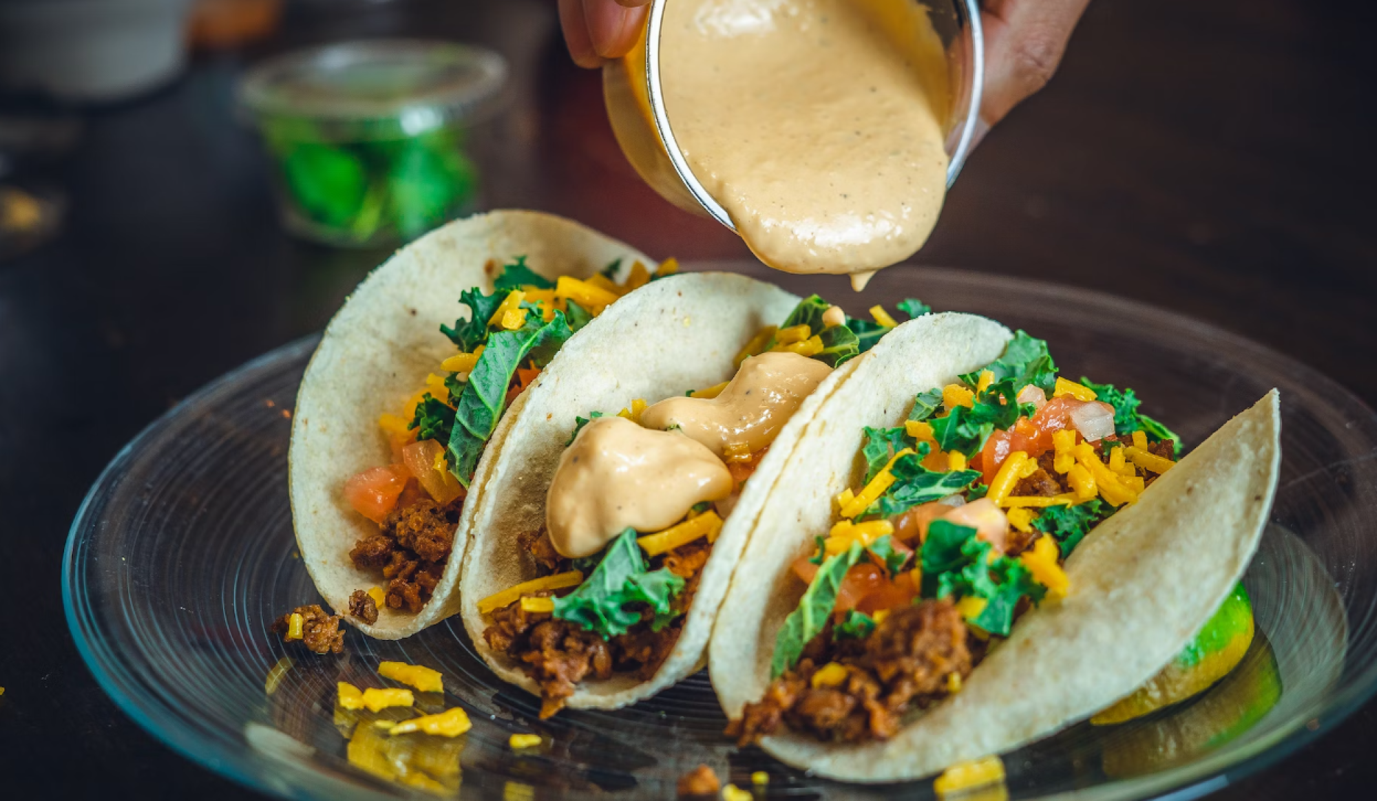 Three tacos on a plate with shredded meat, diced vegetables, and cheese sauce drizzled from a spoon.