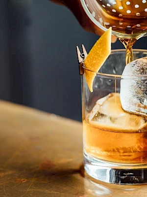 A bartender pours amber whiskey into a rocks glass with ice, topped with an orange slice and toothpick garnish.