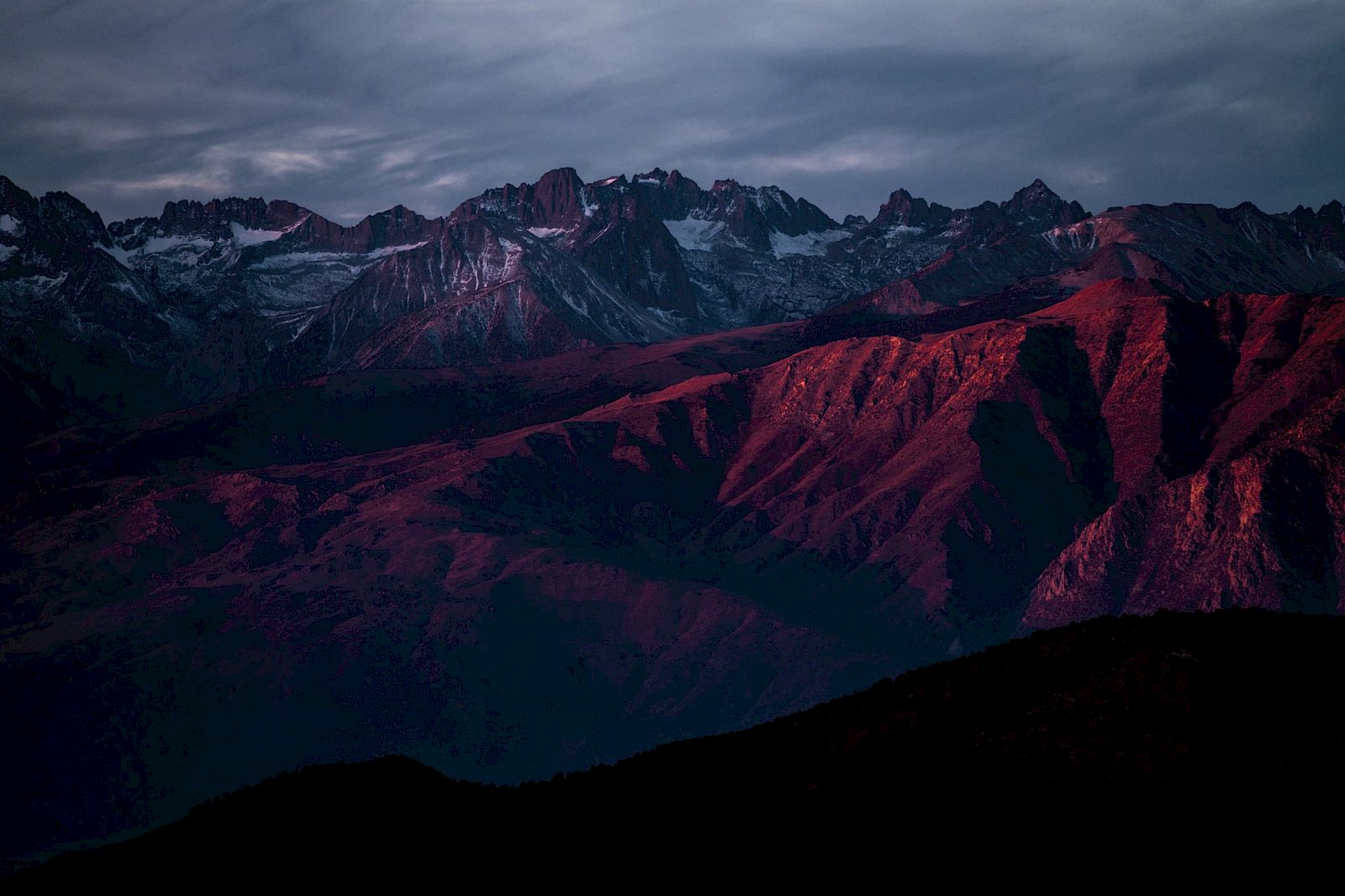A dramatic mountain range at dusk with snowy peaks and red-lit slopes under stormy clouds.