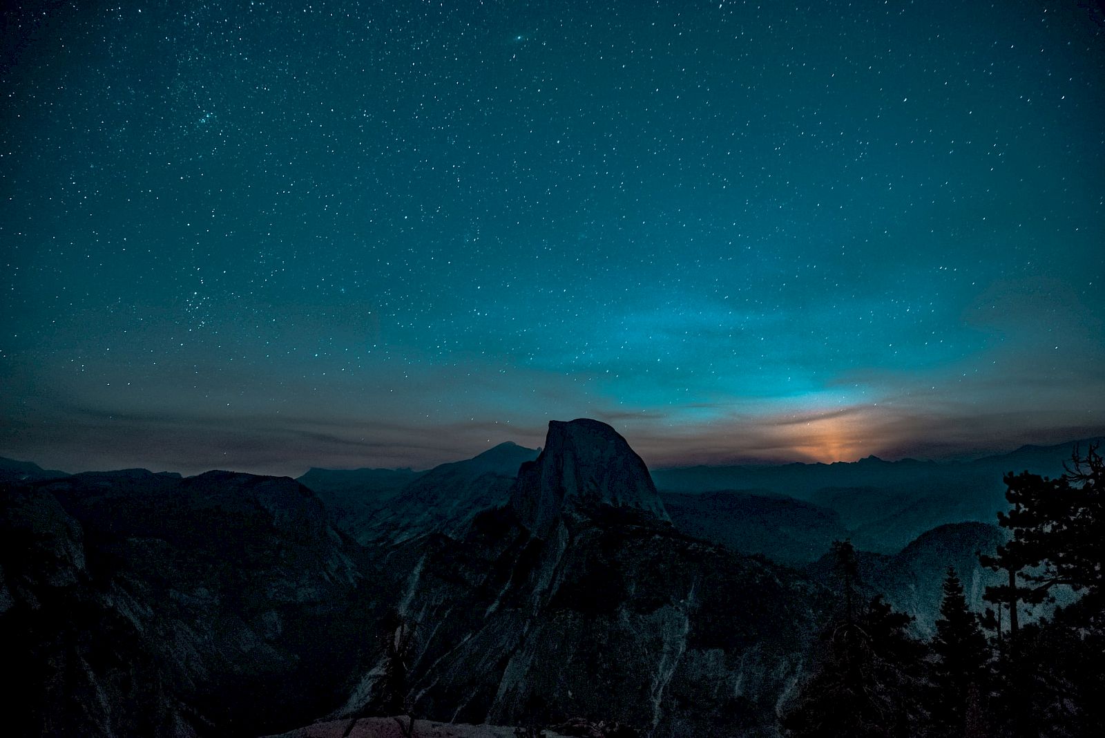 A starry night sky over dark mountain peaks with a glowing horizon and clouds drifting through the valley.