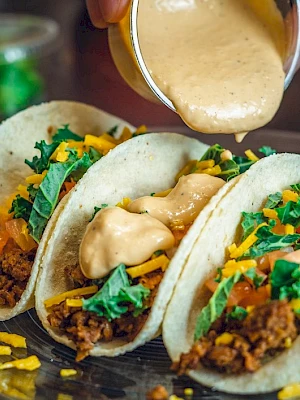 Three tacos on a plate with chopped toppings, with creamy cheese sauce being poured over the top, garnished with herbs and spices.