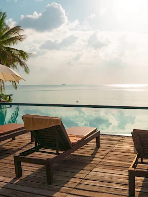 Tropical resort deck by a pool with sun loungers, umbrellas, and a calm ocean horizon under sunny clouds.