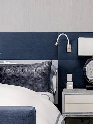 A modern bedroom with a blue upholstered bed, decorative pillows, and a bedside table with lamp and clock.