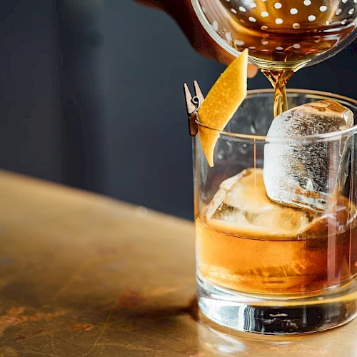 An amber cocktail with ice in a rocks glass is being poured and garnished with a lemon twist, with droplets catching the light.