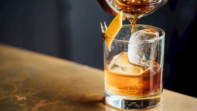 An amber cocktail with ice in a rocks glass is being poured and garnished with a lemon twist, with droplets catching the light.