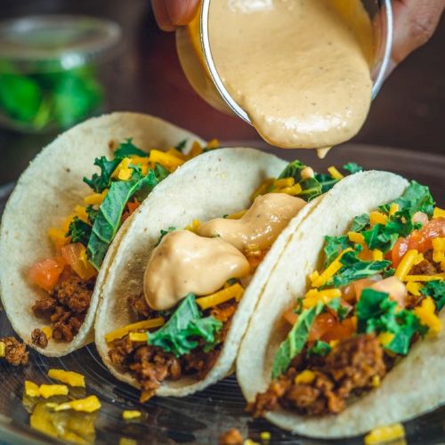 Three tacos on a plate, topped with cheese sauce, chopped cilantro, and crunchy bits in bright colors.
