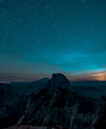 A star-filled night sky glows over dark mountain peaks, with a faint aurora-like green band above drifting clouds.