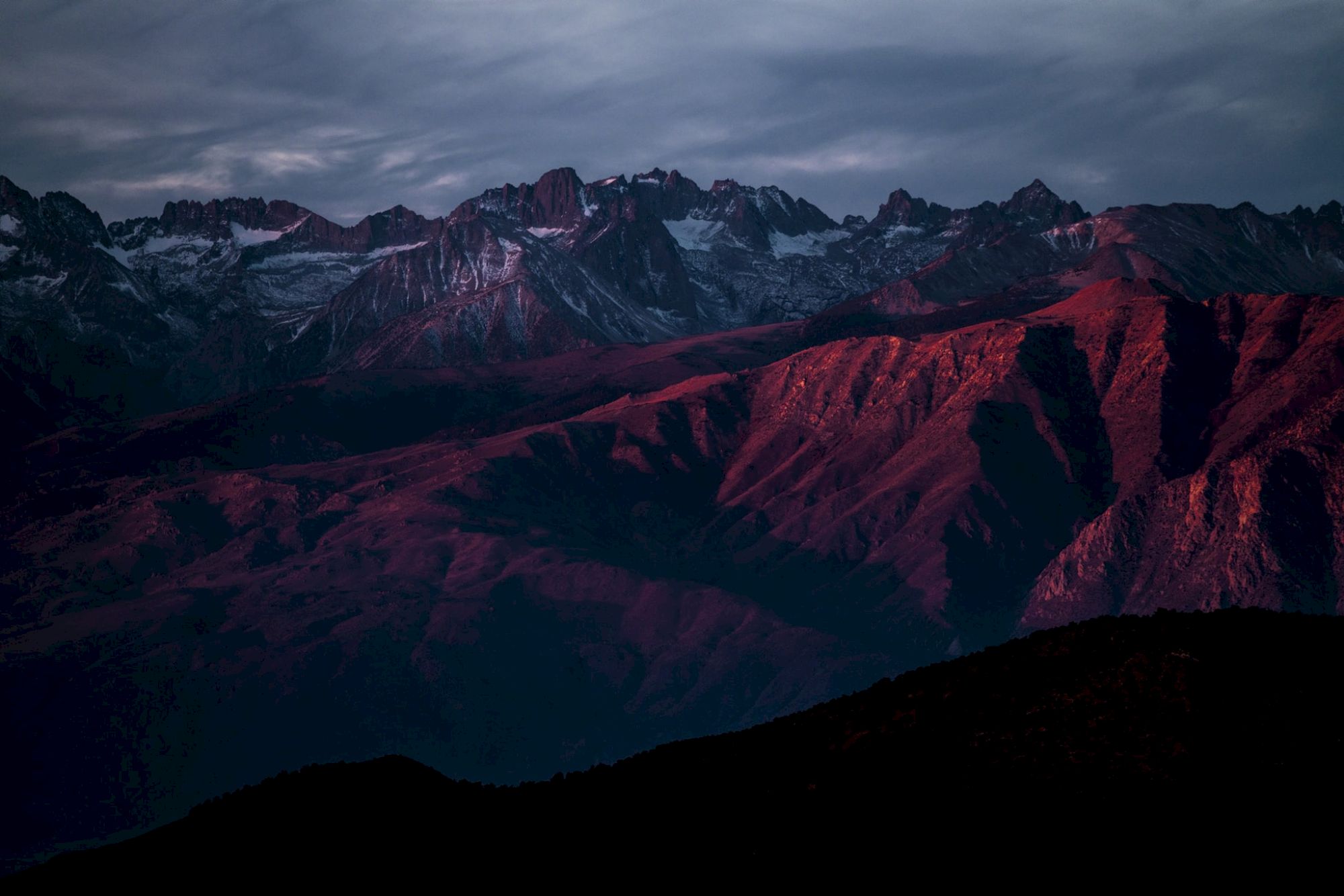 Snowy mountain range at dusk, with rugged peaks and red-lit slopes under a cloudy sky.