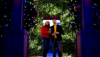 Two people in winter coats stand in a doorway wrapped in glowing green holiday lights, holding warm drinks.