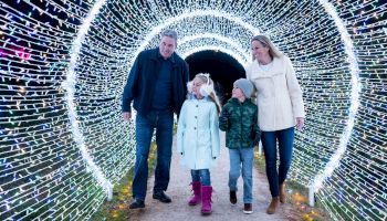 A family walks together inside a glowing holiday light tunnel with colorful string lights, enjoying the festive nighttime atmosphere.