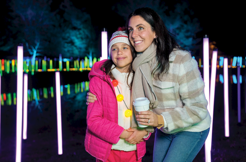 Two women hug at night by colorful glowing light tubes, one in a pink jacket holding a hot drink.
