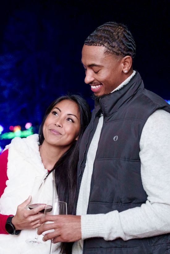 A smiling couple stands close together at night, holding drinks, with colorful city lights blurred in the background.