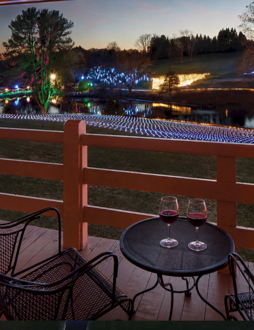 A riverside patio table and chairs look over a pond/stream covered in colorful holiday lights at dusk.