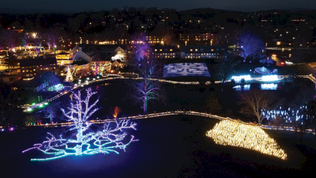 Aerial night view of a landscaped park with buildings, trees lit in blue and purple, and glowing light displays along paths.