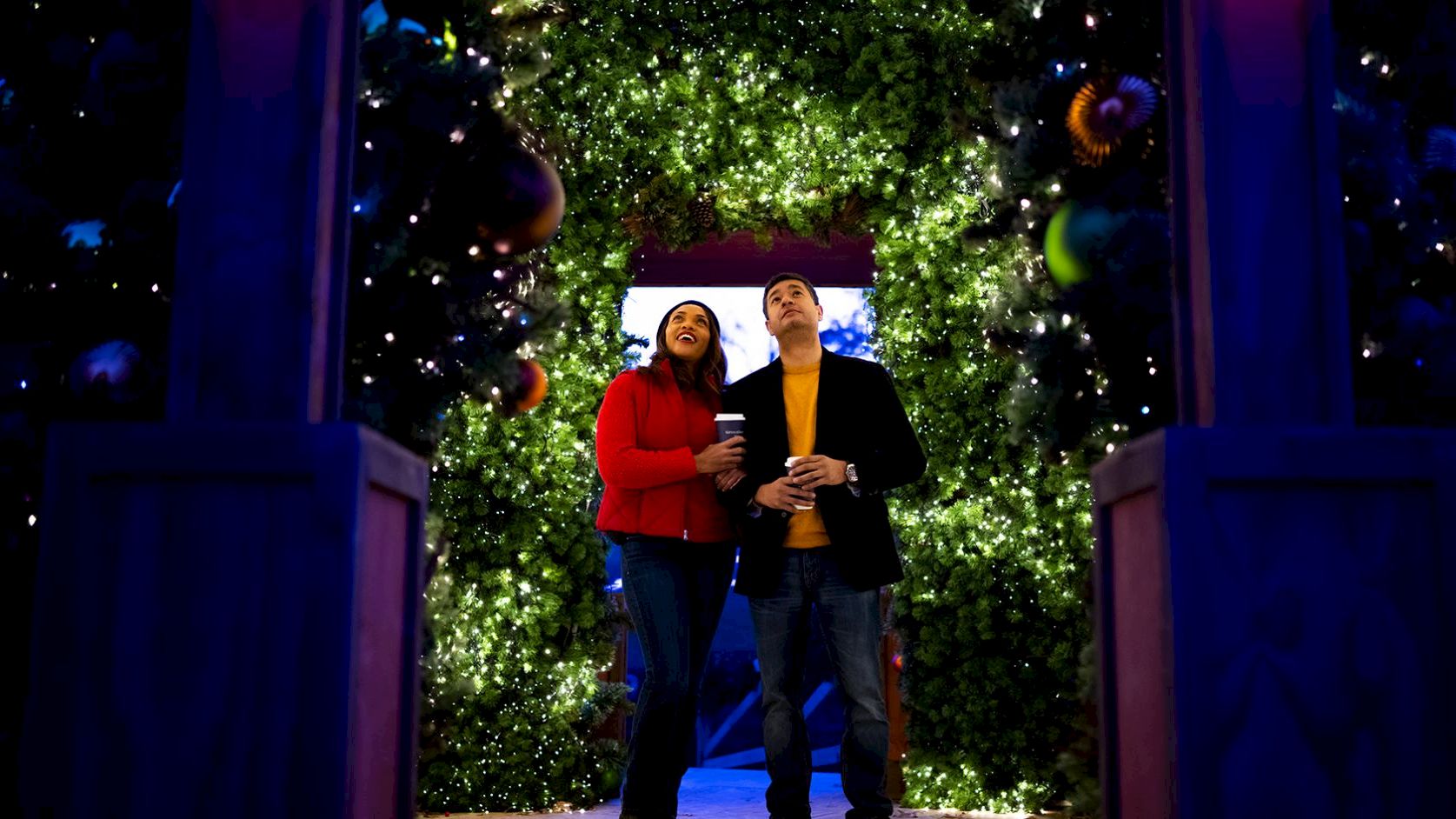 A couple stands under a glowing, tree-lit archway with festive lights and hanging ornaments, holding drinks and smiling at the camera.