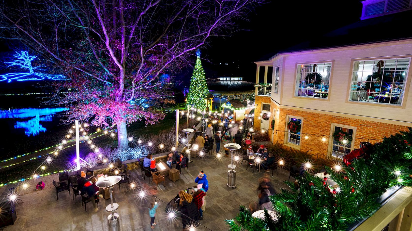 People dining and mingling at an outdoor nighttime venue, string lights glowing, a tall Christmas tree, festive decor, and a brick building lit up in the background.