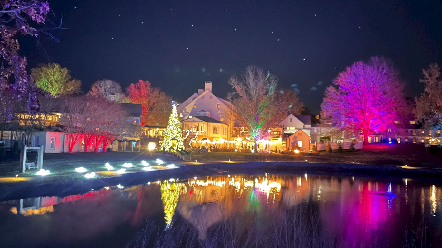 A festive village by a calm pond, with colorful lights, a glowing Christmas tree, and a starry night sky.