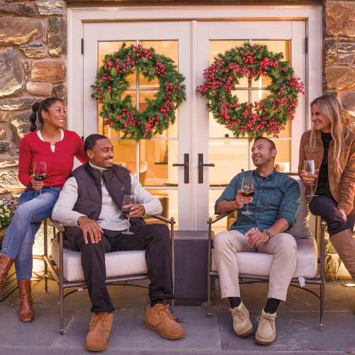 A group of friends lounges on an outdoor patio with Christmas wreaths, holding drinks by a glowing fireplace, smiling in the holiday season.