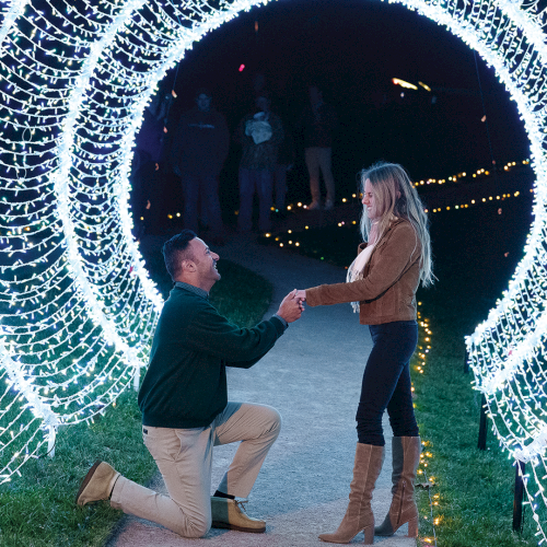 A man kneels to propose to a woman under glowing holiday light tunnels at night.