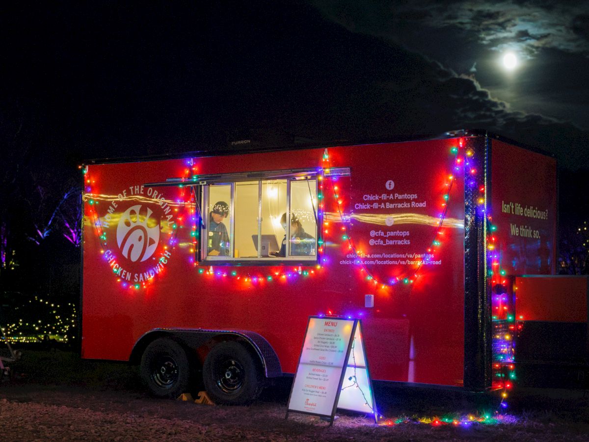 A brightly lit Chick-fil-A food truck at night with colorful string lights and a menu sign outside.