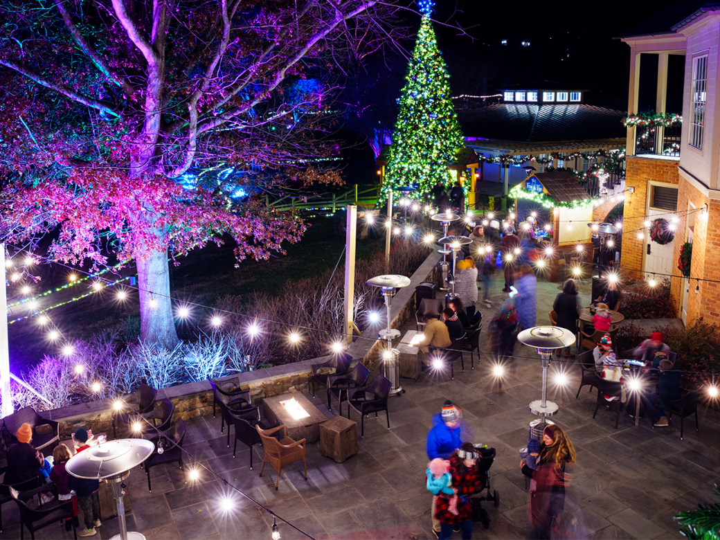 A festive outdoor patio at night with people gathered around firepit heaters, twinkling lights, and a tall decorated Christmas tree.