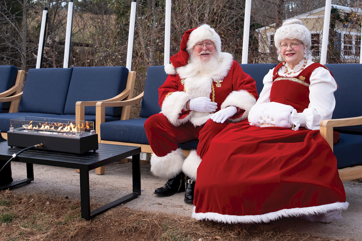 Santa and Mrs. Claus relax outdoors on blue patio chairs beside a glowing fire pit in the winter woods.