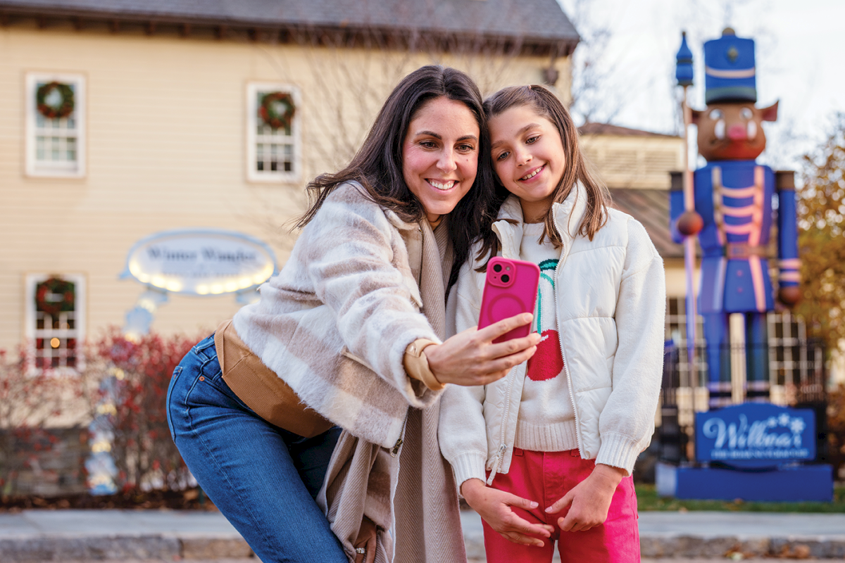 A mom and daughter smile for a photo outdoors by a big blue nutcracker statue and a &ldquo;Welcome&rdquo; building sign.