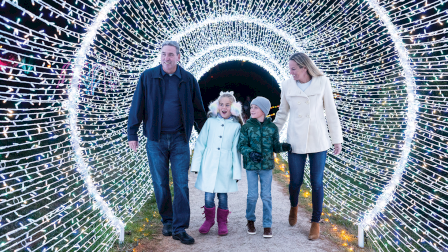 A family strolls through a tunnel of colorful Christmas lights, smiling together on a winter walkway.